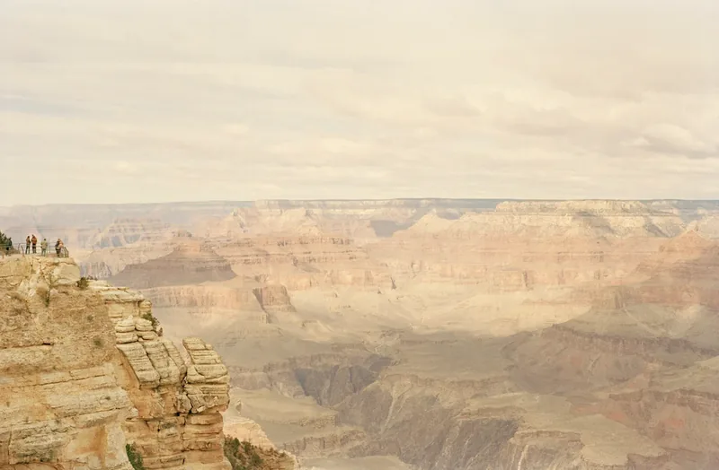 A Grand Canyon — softly hazy photograph of the Grand Canyon's layered sandstone and limestone formations with tiny figures on the rim for scale. Medium format color film by Alex Hoerner.